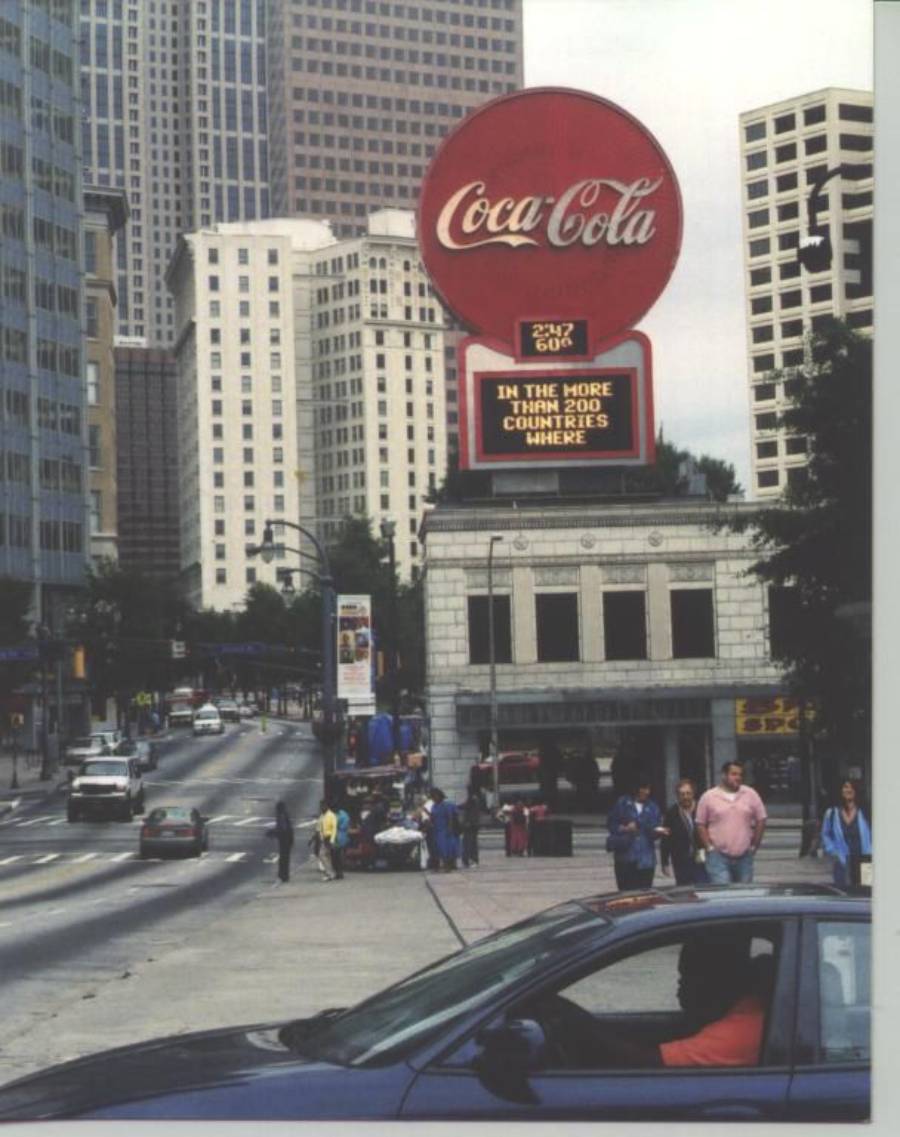 A city street scene with people walking and cars driving by. A large Coca-Cola sign stands above a building, with tall skyscrapers in the background. The Coca-Cola sign reads, "In the more than 200 countries where.