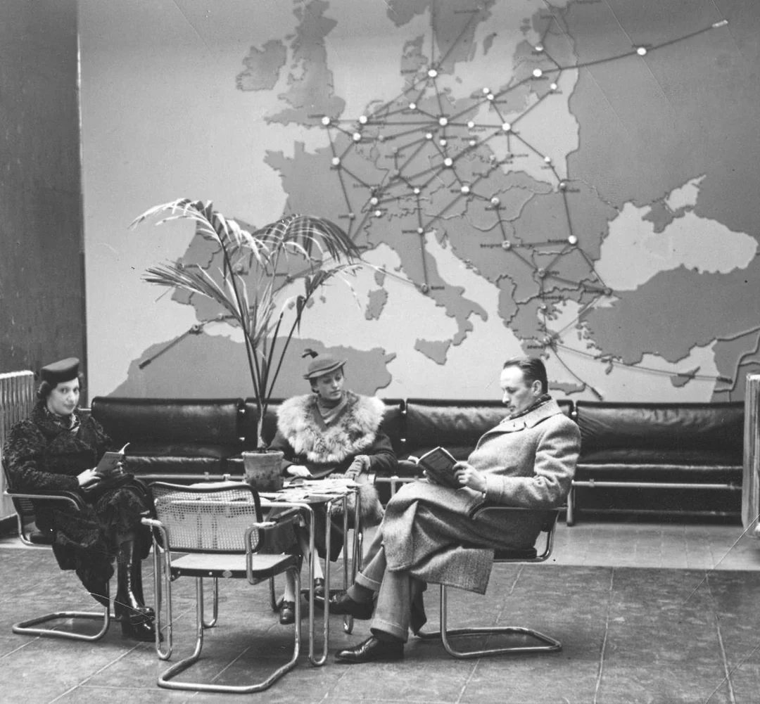 Three people sit reading in a lounge area with modern chairs and a small table. Behind them is a large map of Europe with routes and cities marked, and a tall potted plant is nearby.