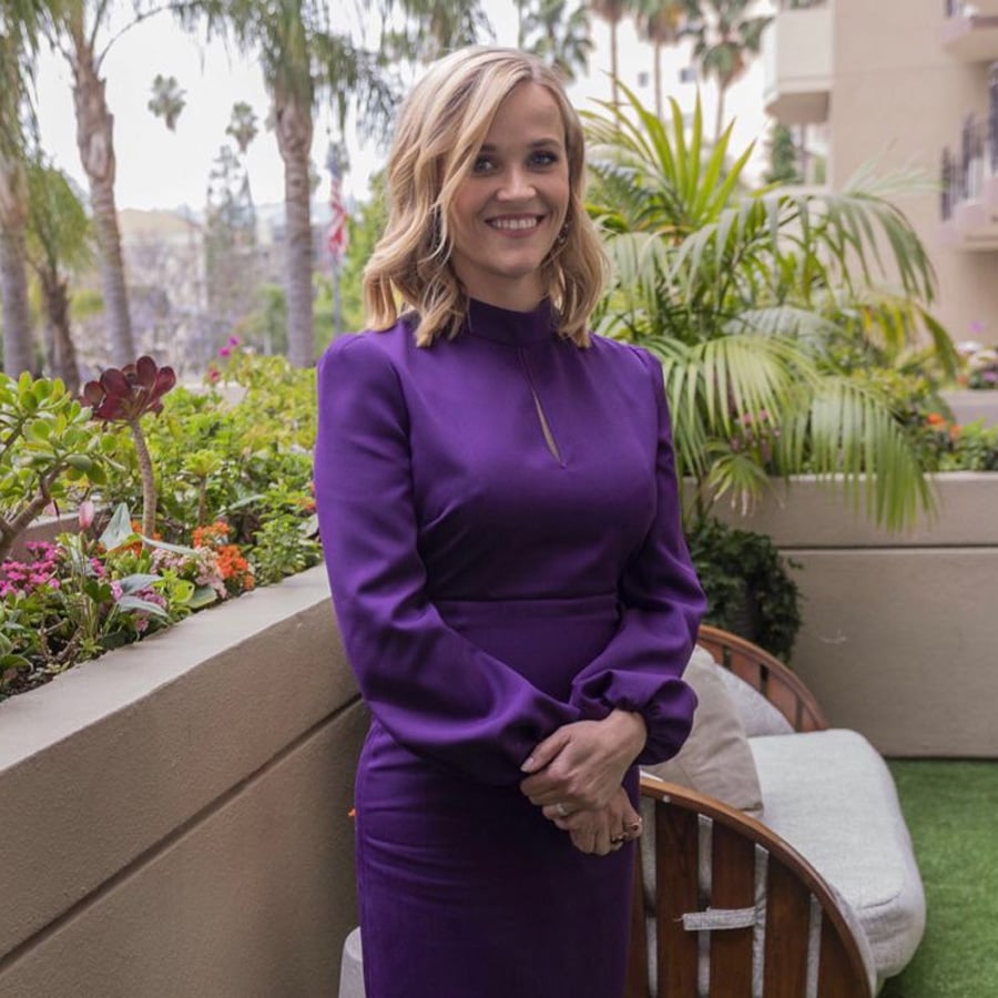 A woman in a long-sleeved purple dress stands outside on a balcony with tropical plants, smiling at the camera.
