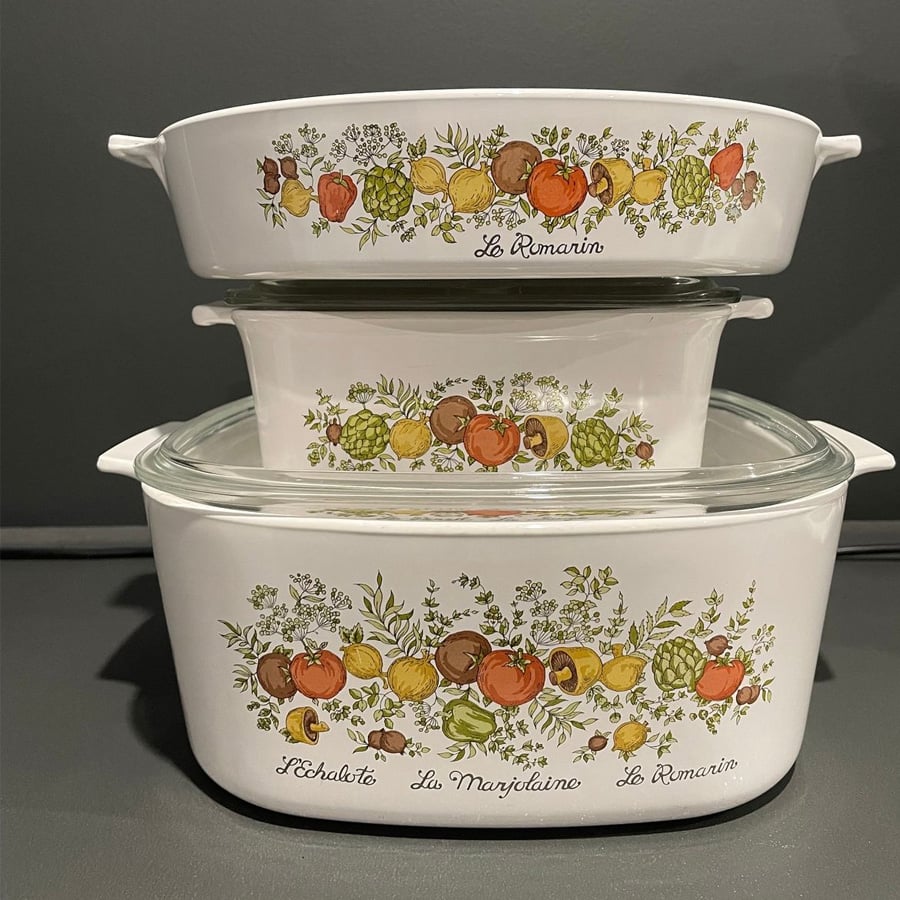 Three vintage white casserole dishes with clear lids, stacked on top of each other, decorated with colorful illustrations of vegetables and herbs, and French text labels like "Le Romarin" and "L’Echalote.