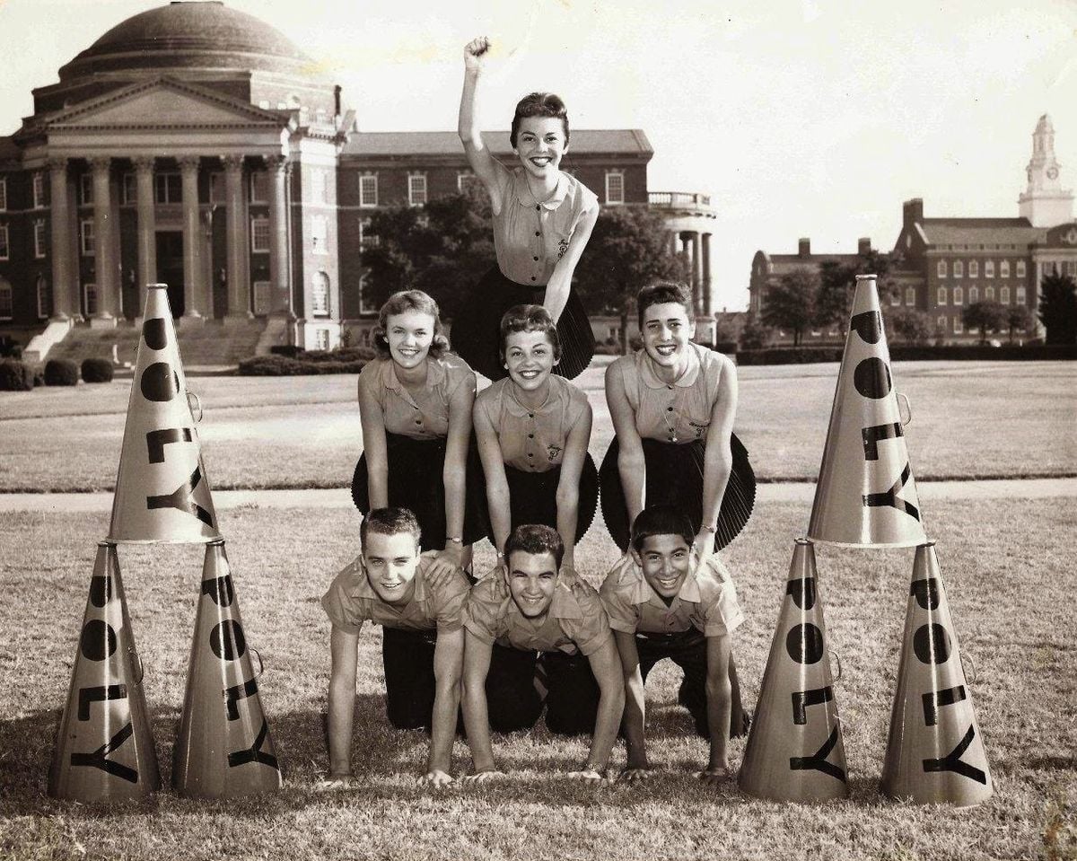 Six cheerleaders form a human pyramid on grass, smiling, with large megaphones and a domed building in the background. One girl stands on top, raising her arm in the air.
