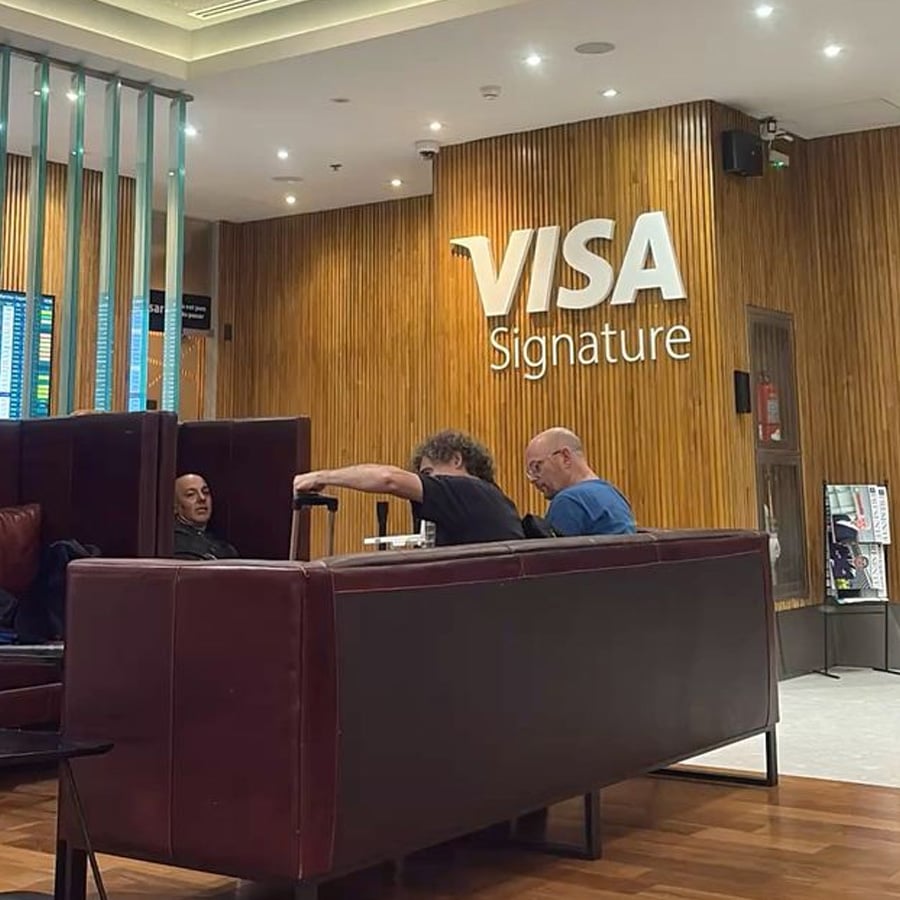 Three people sit on maroon couches in a modern lounge with wood-paneled walls. A large "VISA Signature" sign is displayed on the wall behind them. Bright ceiling lights illuminate the area.
