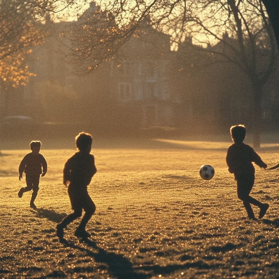 Three children play soccer on a grassy field at sunset, with long shadows cast by the low sun and trees visible in the background.