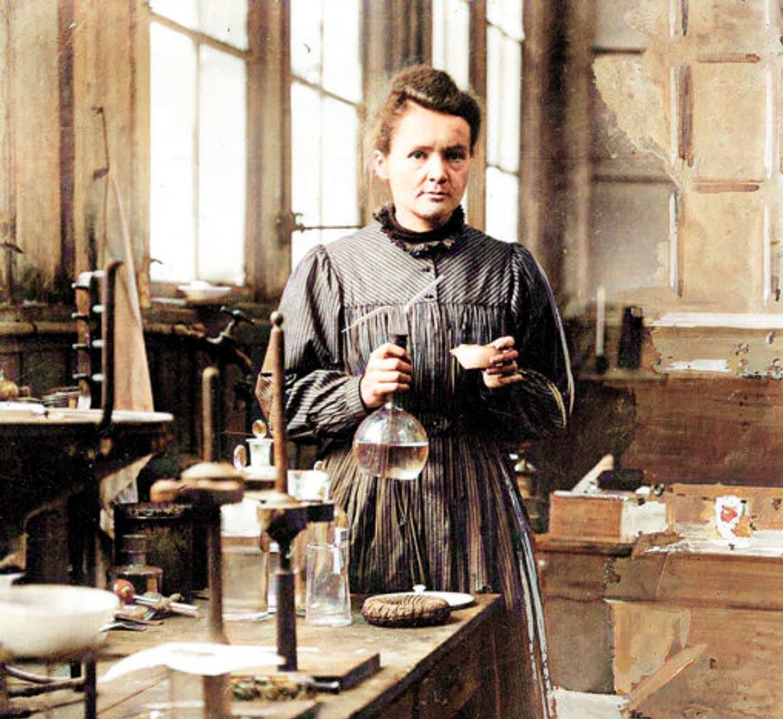 A woman in a striped dress stands in a laboratory, holding a flask and a small cup, surrounded by scientific equipment and wooden tables.