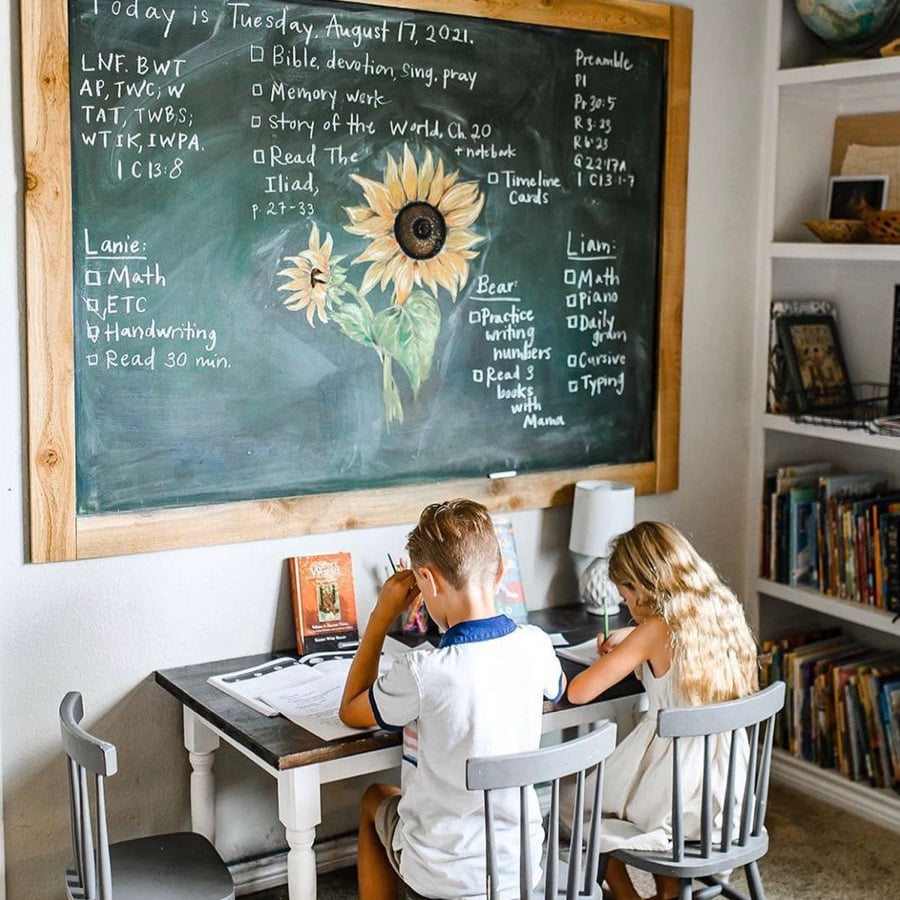 Two children sit at a desk doing schoolwork in a cozy room. Behind them is a large chalkboard with sunflowers and a handwritten lesson plan. Bookshelves filled with books are visible on the right.