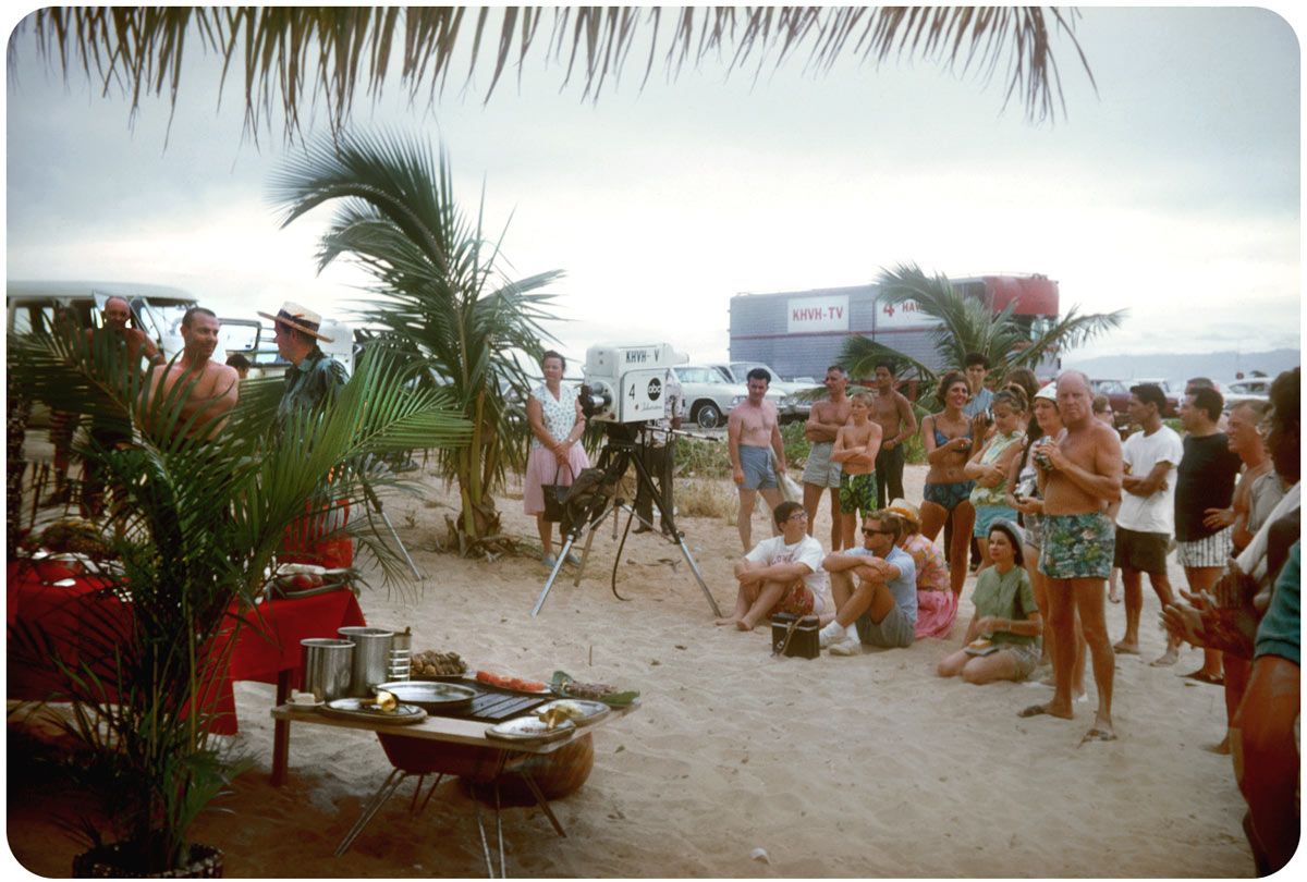 A group of people, some in swimwear, gather on a sandy beach near palm trees and a film camera. Food is set out on tables, and TV equipment and vehicles are visible in the background.