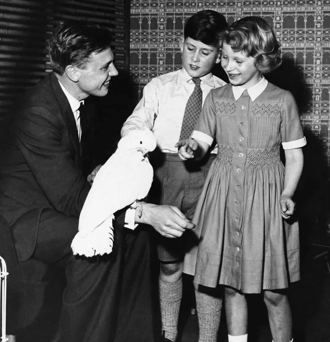 A man in a suit holds a white cockatoo while a young boy and girl smile and reach out toward the bird. The children look excited, standing in front of a patterned wallpaper.