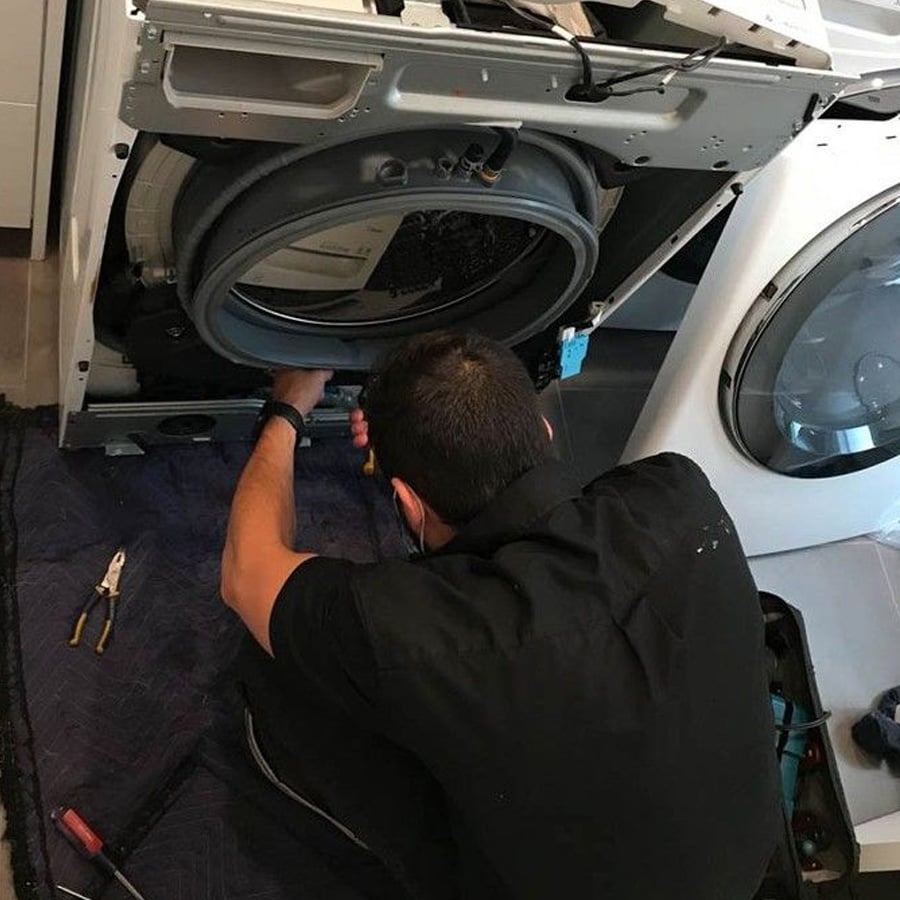 A person kneels on a mat while repairing the inside of a washing machine using tools. Another laundry machine is nearby.
