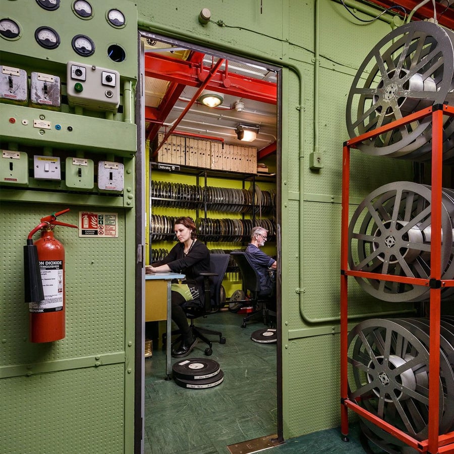 Two people work at desks in a film archive room filled with shelves of film reels. Large reels are stored on racks and tables; a fire extinguisher is mounted on the wall near electrical switches.