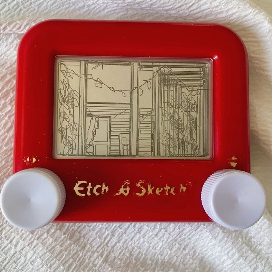 A red Etch A Sketch displays a detailed line drawing of a porch with string lights, posts, and plants. The toy rests on a textured white surface.