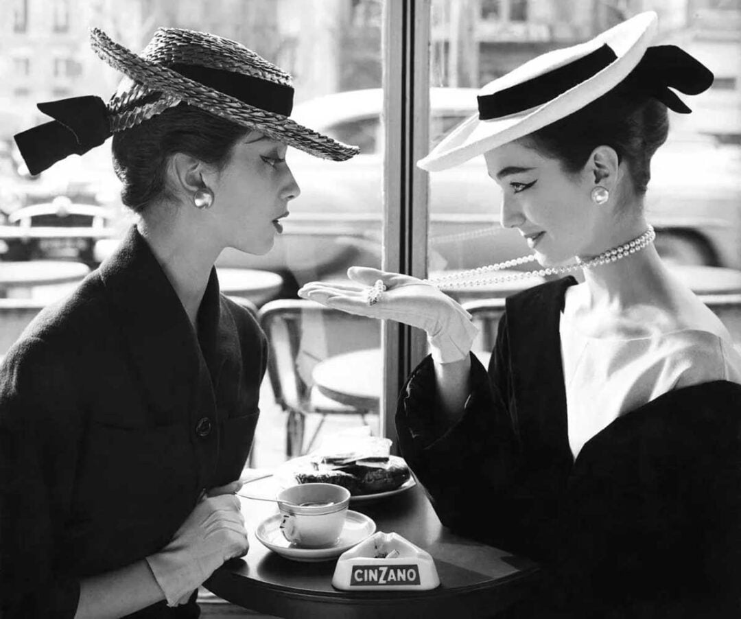 Two elegant women in hats and gloves sit at a café table. One woman gestures with her gloved hand, showing off a ring, while the other looks on. A Cinzano sign and tea cup sit on the table.