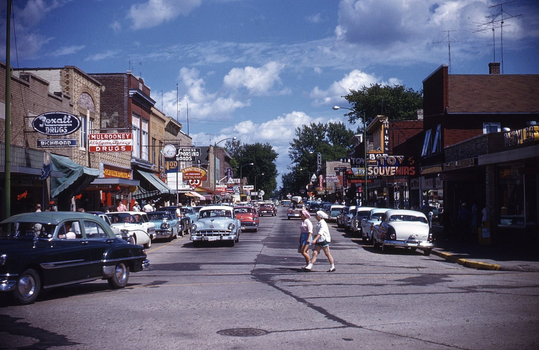 A busy 1950s street scene with vintage cars parked on both sides, people crossing the road, and storefronts with colorful signs, including drugstores and diners, under a partly cloudy sky.