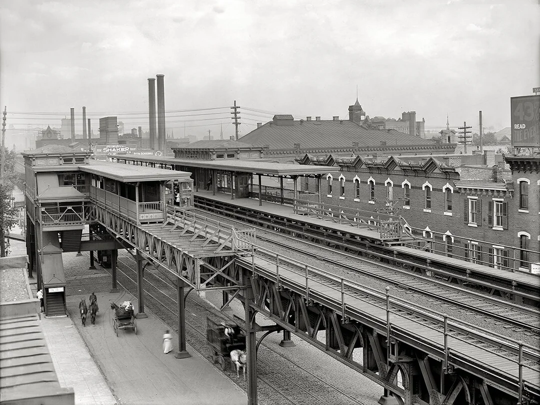 Black and white photo of an elevated train station above a city street with vintage cars, pedestrians, brick buildings, and factory chimneys in the background.