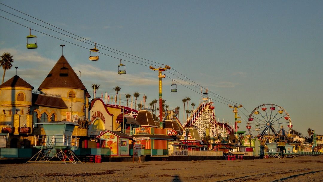 Colorful amusement park by the beach at sunset, featuring rides like a Ferris wheel, roller coaster, cable car ride, and whimsical buildings, with palm trees in the background and empty sandy foreground.