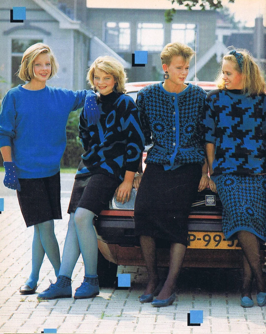 Four women in blue and black outfits lean against a red car, smiling. The background shows an unfinished building. The women wear patterned sweaters, skirts, and matching accessories. The scene has an 80s fashion vibe.