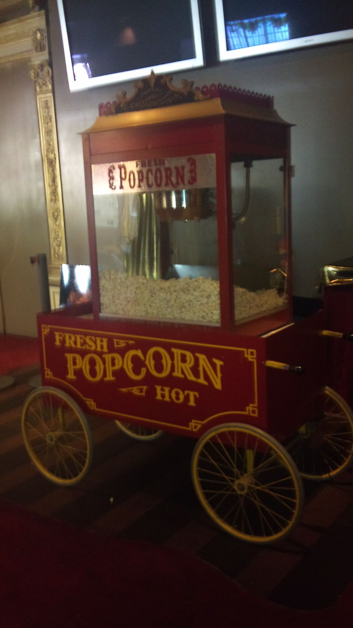A vintage-style red popcorn cart with gold trim and "Fresh Popcorn Hot" written on the side is filled with popcorn and stands on a carpeted floor indoors.
