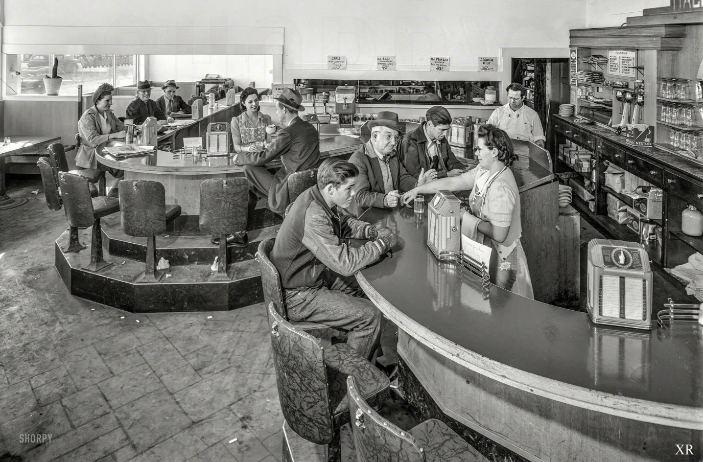 Black and white photo of people sitting at a curved diner counter, being served by a waitress. Other diners and staff are visible in the background; shelves with goods line the walls. The scene appears to be from the 1940s.