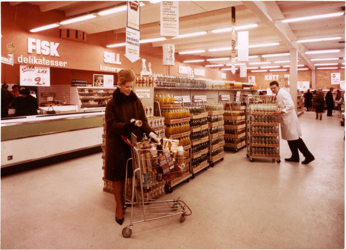 A woman in a dark coat shops in a grocery store, placing items in a cart. A man in a white coat stands near shelves filled with bottles. Signs with Swedish text hang from the ceiling. The store has bright lighting and wide aisles.