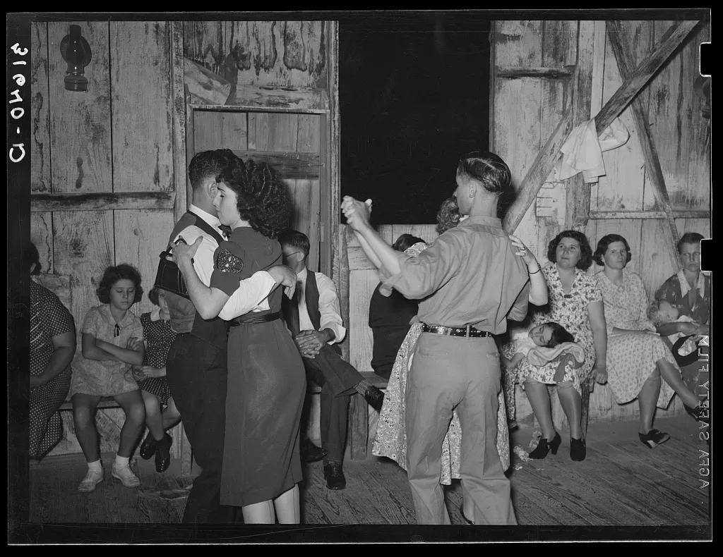 Couples dance closely on a wooden floor while several women sit along the wall, some watching and others talking, in a rustic, wooden room.