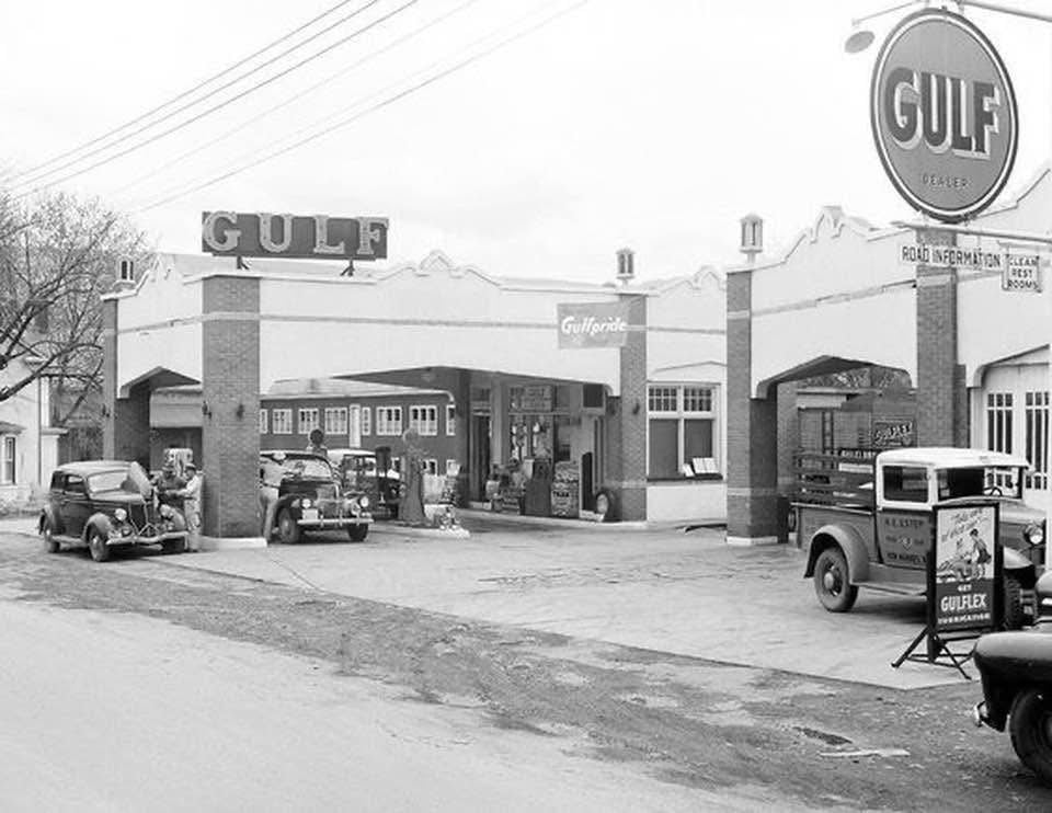 Black and white photo of a vintage Gulf gas station with old cars and trucks parked outside, a large Gulf sign above the building, and people near the pumps. The scene appears to be from the early to mid-20th century.