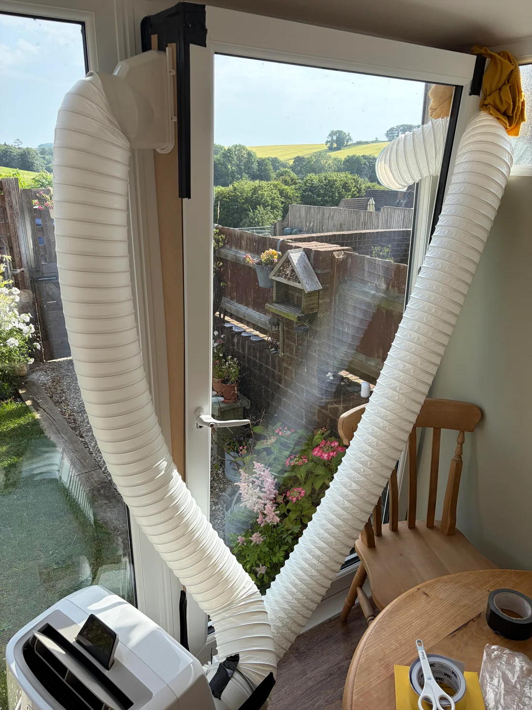 Two large white exhaust hoses are connected from a portable air conditioner inside to a window and door, both partly open and sealed with foam. A wooden chair and garden outside are visible.