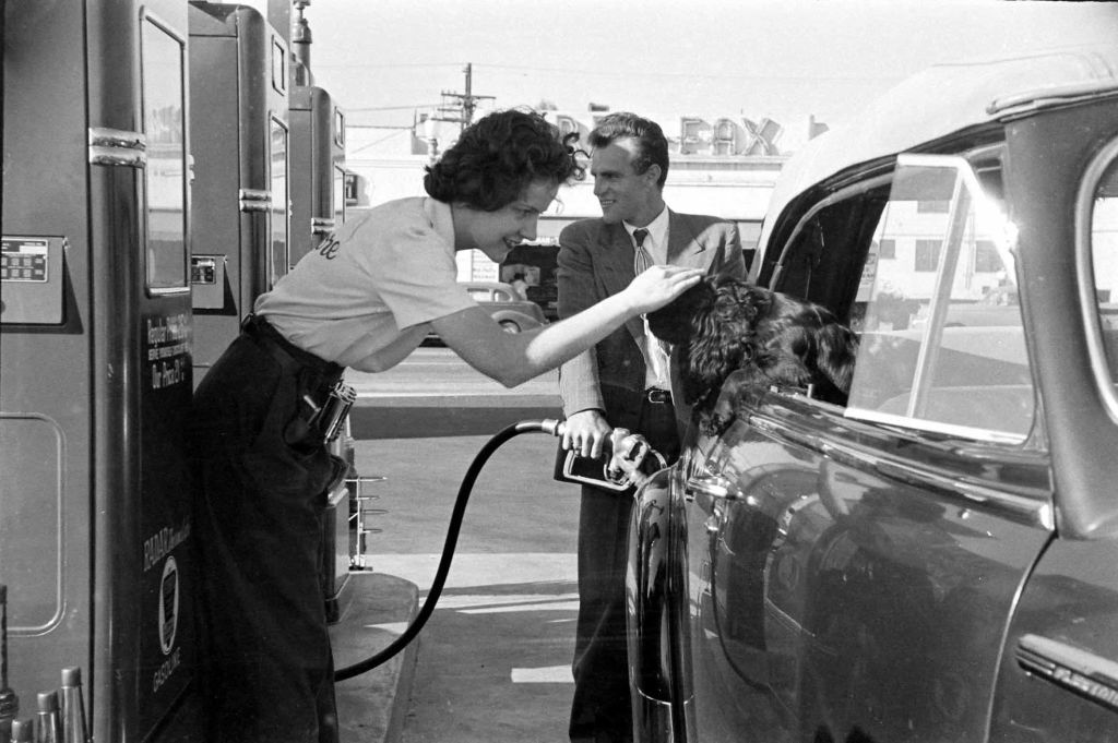 A woman pumps gas while petting a dog in the back seat of a car. A man stands nearby, smiling. The scene appears to be from a vintage gas station.
