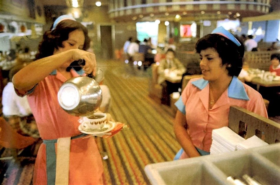 Two waitresses in pink uniforms work in a busy restaurant; one pours coffee into a cup on a saucer, while the other stands nearby. Diners sit at tables in the background.