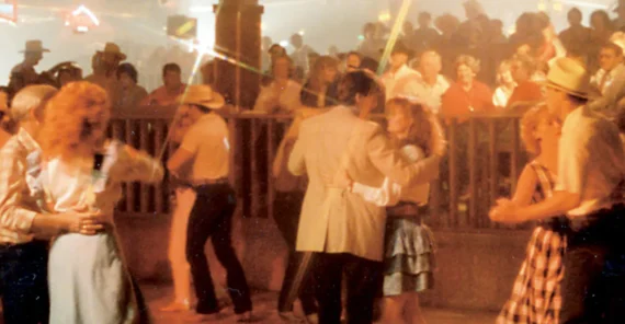 People in cowboy hats and western outfits dance under colorful lights in a crowded, lively country-western bar or dance hall, with a cheering audience in the background.