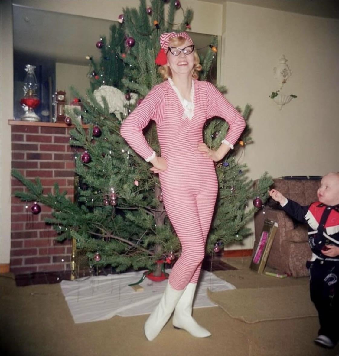A woman in pink striped pajamas and white boots poses happily by a Christmas tree, while a small child in a dark outfit stands nearby, pointing. The room is decorated for the holidays.
