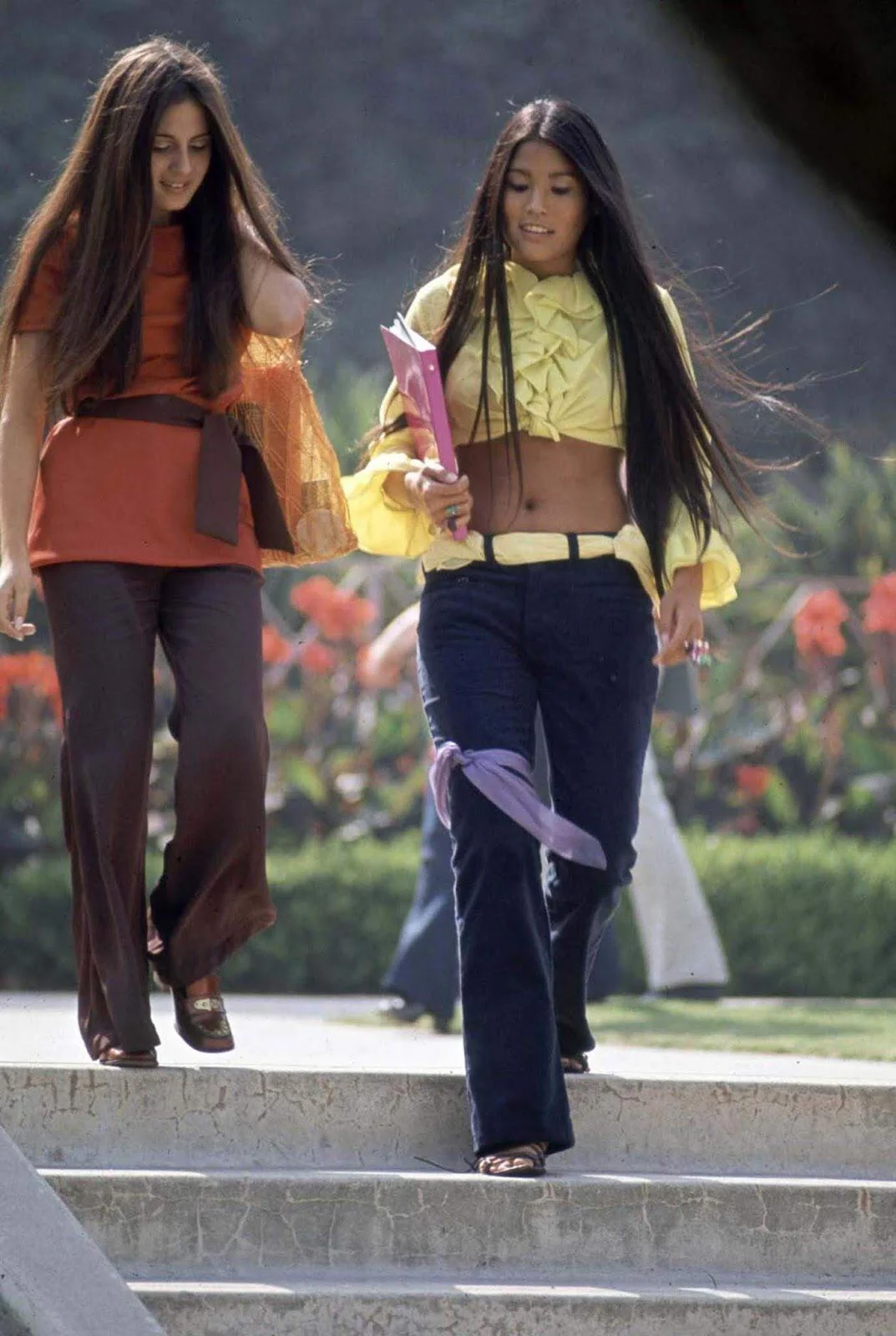 Two young women with long hair walk down steps outdoors. One wears a burnt orange top and brown flared pants; the other wears a yellow ruffled crop top and dark flared jeans, holding books and papers. Flower beds are in the background.