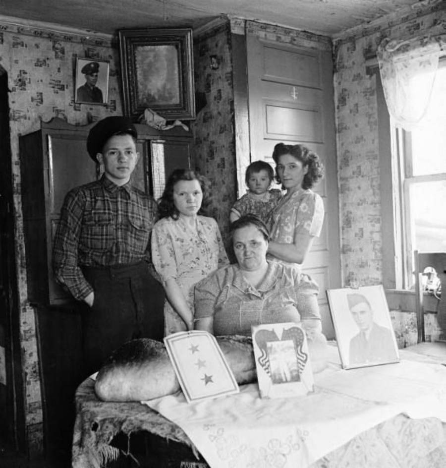 A family of five poses in a modest room. Three women, a girl, and a boy stand behind a table displaying two large loaves of bread and framed photos of men, likely soldiers. A military portrait is on the wall above them.