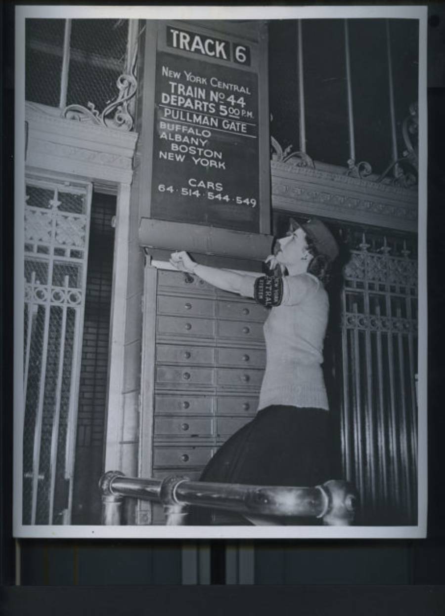 A woman stands at a train station under a sign for Track 6, showing destinations like Albany and New York, reaching up towards a set of lockboxes. The photo appears to be from a past era.