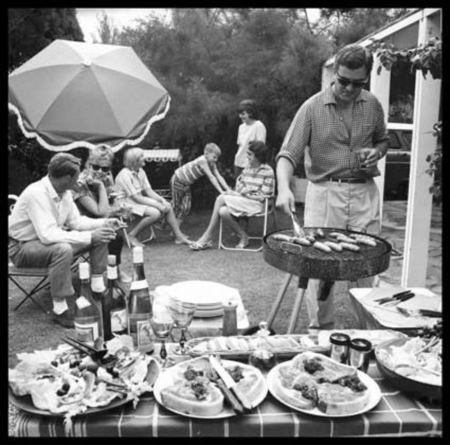 A man grills food beside a table filled with plates and drinks at an outdoor gathering. Several people sit and talk in the background near a large umbrella on a sunny day.