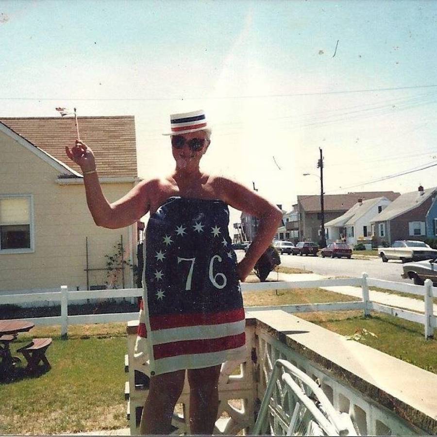 A person in a patriotic outfit with "76" on it stands outside, holding a cigarette and wearing sunglasses and a festive hat, with houses and a yard in the background.