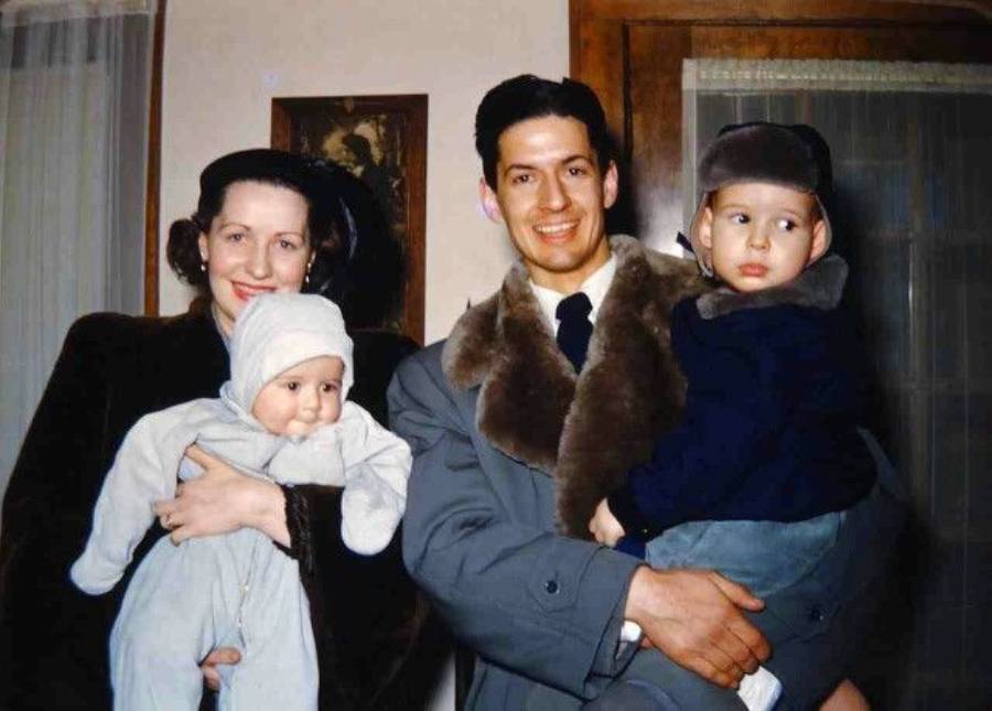 A woman and man, both in vintage winter clothing, smile while holding two young children, one dressed in white and the other in dark blue, inside a warmly lit room.