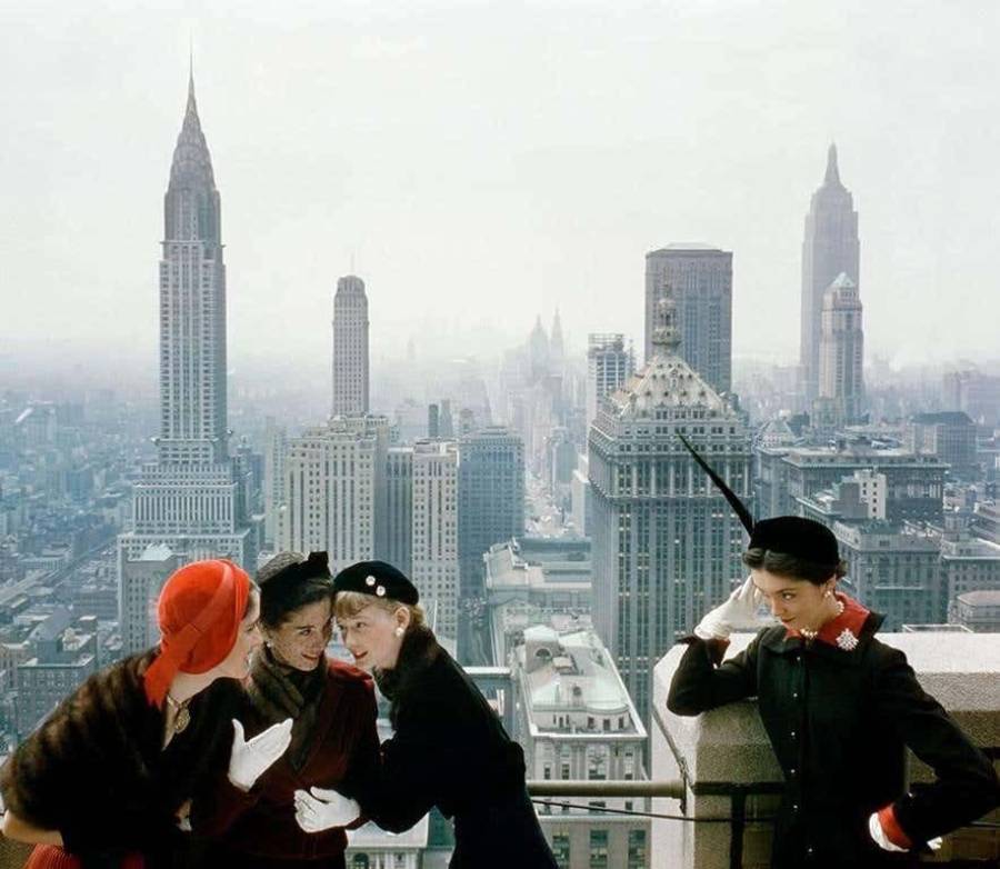 Four women in vintage clothing pose on a rooftop with the New York City skyline, including the Chrysler and Empire State Buildings, in the background. Two women appear to be whispering to a third.