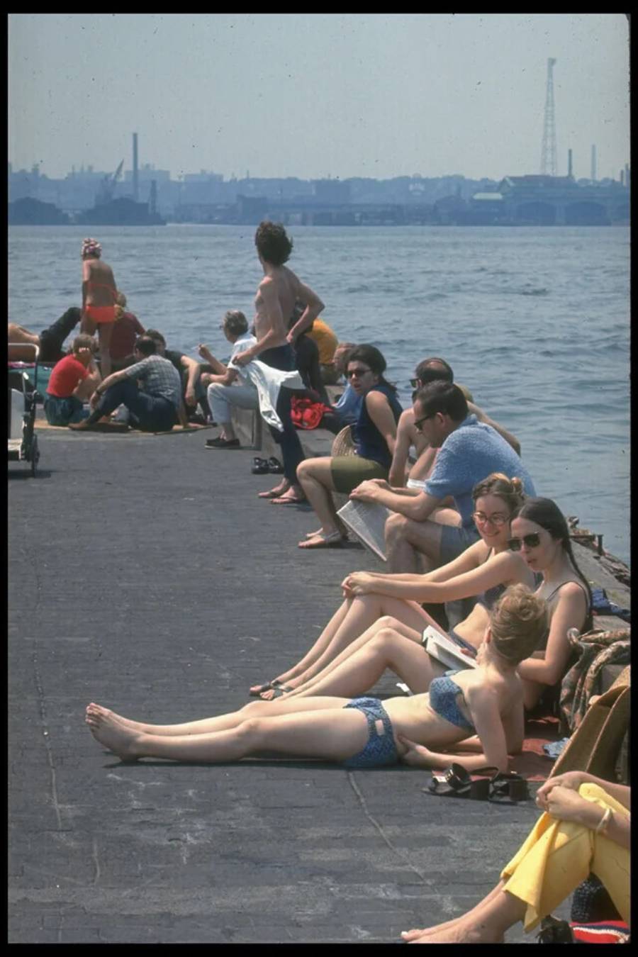 People in swimsuits relax and sunbathe on a pier by the water, some sitting and some lying down. The city skyline and industrial structures are visible across the water on a hazy day.
