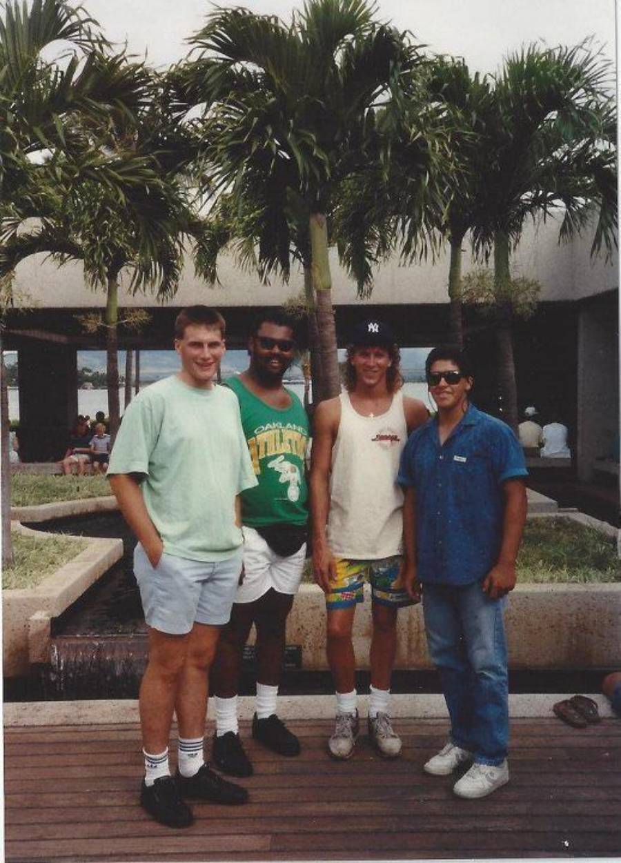 Four men stand together outdoors near palm trees. They are casually dressed in shorts and t-shirts, smiling at the camera. The background features a modern building and people sitting by a water feature.