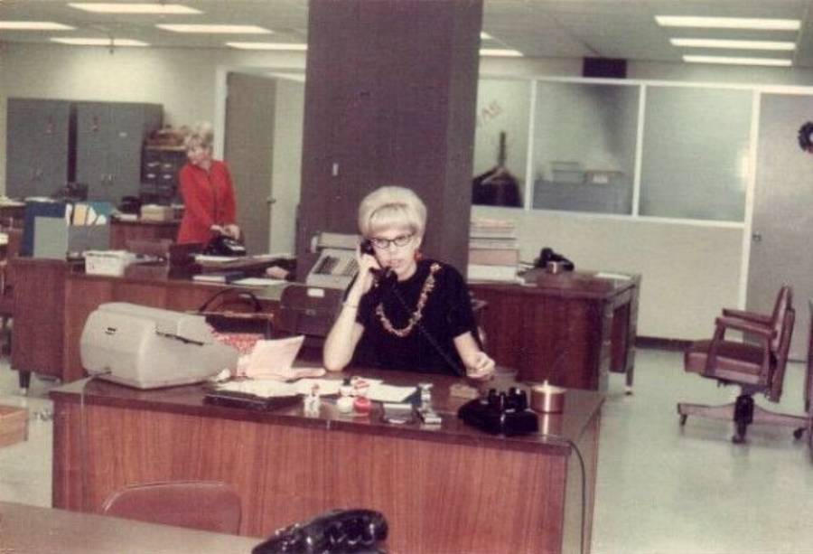 A woman with short blonde hair and glasses sits at a desk in a vintage office, talking on a corded phone. Another woman stands in the background near filing cabinets. The room has typewriters and office supplies.