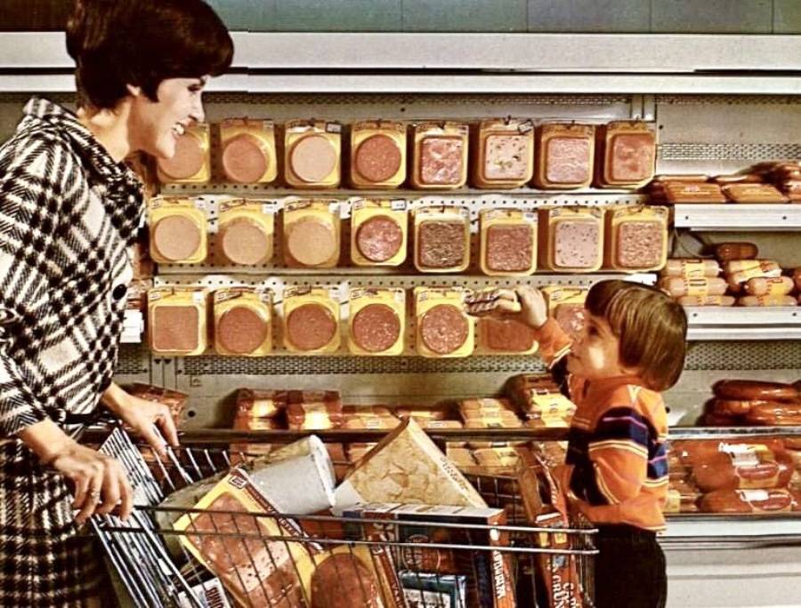 A woman and a young child stand in a grocery store meat aisle. The woman smiles at the child, who is pointing at packaged meat on the shelves. Their shopping cart is filled with groceries.