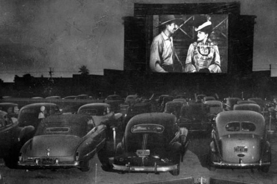 Black and white photo of a drive-in theater at night, with vintage cars parked facing a large outdoor movie screen showing a scene of a man and woman in period costumes speaking to each other.