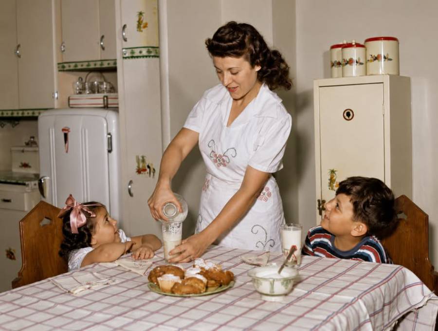 A woman pours milk for two children seated at a kitchen table with pastries. The kitchen has vintage decor, including a white refrigerator and yellow cabinets.