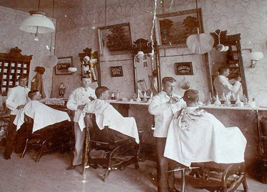 Sepia-toned photo of an old-fashioned barbershop with four barbers cutting hair for male clients, mirrors on the wall, and vintage decor.