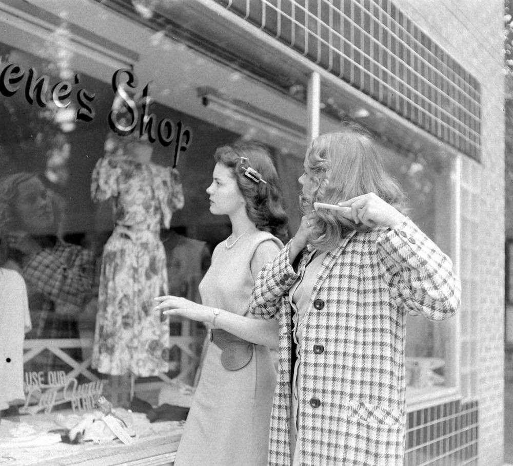 Two young women look into a clothing shop window display. One wears a plaid coat and gestures with her hand, while the other stands beside her in a light dress. Dresses and shop signs are visible through the glass.