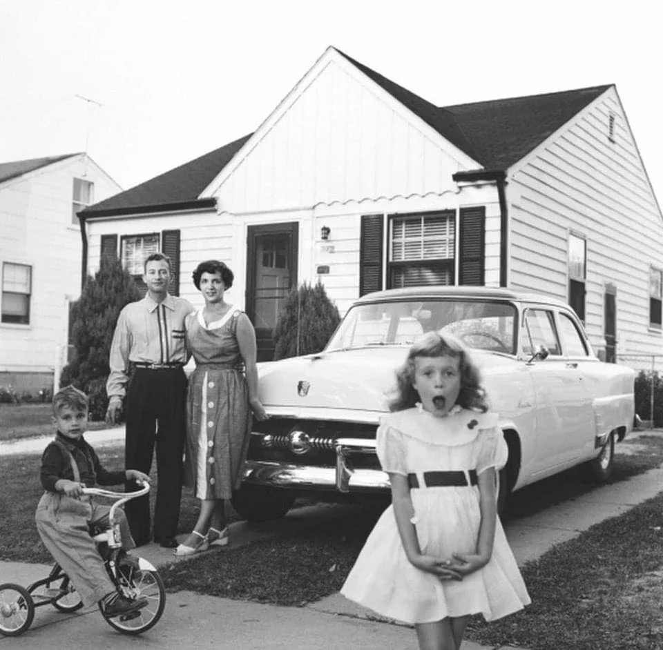 A black-and-white photo of a family in front of a house: a man and woman stand near a classic car, a young boy sits on a tricycle, and a girl in a dress stands closer to the camera looking surprised.