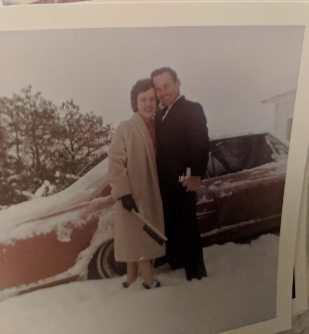 A smiling woman and man in winter coats stand in snow next to a red, snow-covered car. The woman holds a scraper, and both appear happy. Trees and a building are visible in the snowy background.