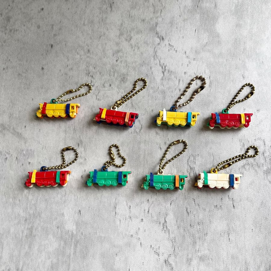 Eight colorful miniature train keychains, each with a gold chain, are arranged in two rows on a gray surface. The trains are in combinations of red, yellow, green, blue, and white.