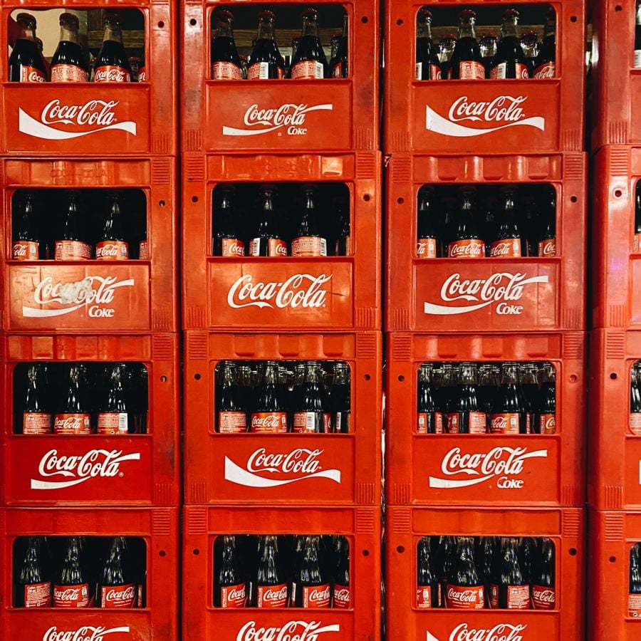 Red plastic crates stacked with glass Coca-Cola bottles. Each crate has the Coca-Cola logo printed in white on the front. The bottles are arranged in rows inside the crates.