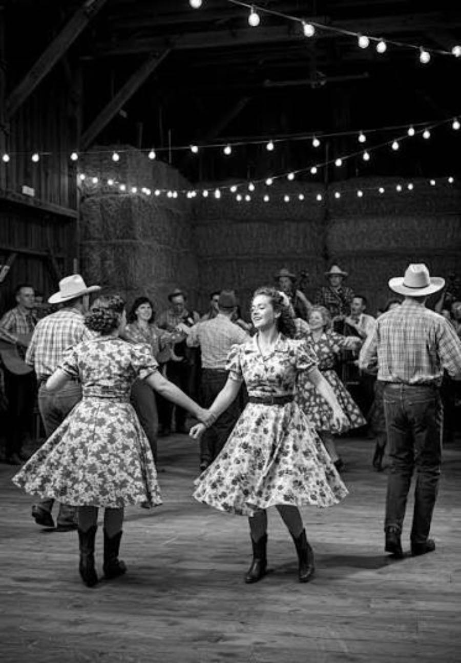 Two women in floral dresses and boots dance in a barn decorated with string lights. People in plaid shirts, some wearing cowboy hats, play music and watch in the background. The scene is lively and festive.