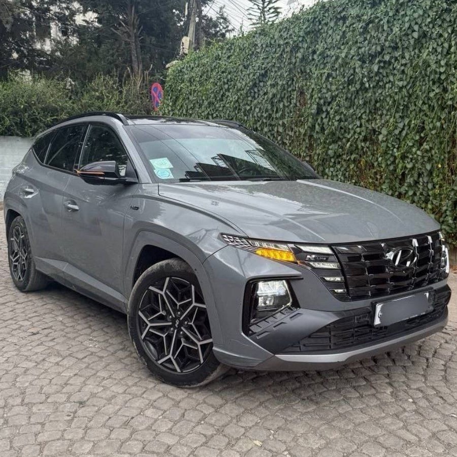 A gray Hyundai Tucson SUV is parked on a cobblestone driveway next to a tall green ivy-covered wall, with some trees and electric wires visible in the background.